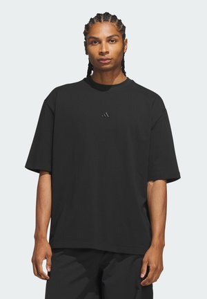 Osoba z warkoczami, ubrana w luźną czarną koszulkę z małym logo Adidas na środku, stojąca na jednolitym tle.