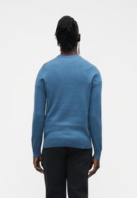 Svetlo modra dolga pulover z dolgimi rokavi, izdelan iz mehke tkanine, z okroglim izrezom in rebrastim robom, nošen s črnimi hlačami, prikazan od zadaj.