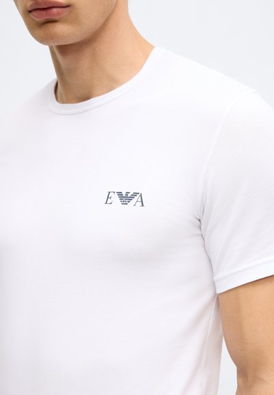 Balts kokvilnas T-krekls ar apaļu kaklu, īsām piedurknēm un mazu zilu logotipu "EVA" kreisajā krūšu daļā. Gluda tekstūra, piegulošs dizains.