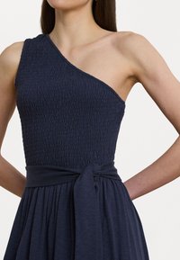 Epäsymmetrinen tummansininen mekko, jossa on teksturoitu smokkibodice, leveä vyötärösolki ja laskostettu, virtaava hame. Sujuva, kevyt kangas.