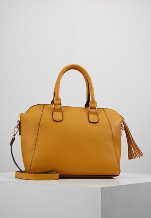 Musztardowożółta teksturowana skórzana torebka z podwójnymi uchwytami, odpinanym paskiem na ramię oraz detalem w postaci frędzla na szarym tle.