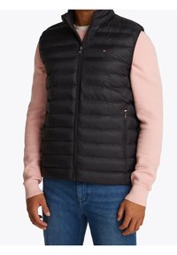 Czarna kamizelka puchowa z przednim zamkiem, z poziomymi przeszyciami i małym logo. Noszona na jasno różowej koszulce z długim rękawem.