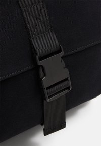 Sac en toile noire avec une surface texturée, équipé d'une fermeture à boucle en plastique et d'une bandoulière réglable, mettant l'accent sur la durabilité et la fonctionnalité.