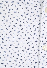 Weiße Bluse mit einem blauen Blumenmuster, verziert mit kleinen Blumen und Blättern. Inklusive eines Knopfdetails im Kragenbereich. Glatte Textur.