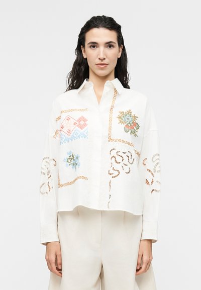 Femme portant une chemise blanche à boutons ornée de motifs floraux et géométriques brodés colorés, associée à un pantalon large beige clair.