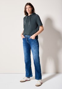 Camiseta de manga corta con capucha en tejido verde oscuro, combinada con jeans rectos azules y zapatillas beigas con acentos rojos. Textura suave.