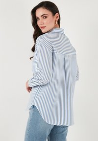 Gestreifte hellblaue und weiße Bluse mit langen Ärmeln, einem Kragen und einem abgerundeten Saum. Der Stoff wirkt leicht und hat eine glatte Textur.