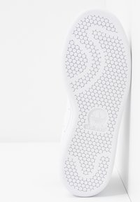 La suela de la zapatilla blanca presenta un patrón de textura hexagonal, con un logotipo destacado en el centro y un diseño de borde suave.