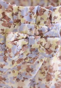 Tissu à motif floral avec des teintes bleu clair, jaune et marron. Présente une taille froncée et un nœud accentué.
