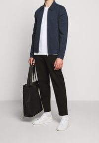 Veste zippée bleu marine, t-shirt blanc, pantalon noir fuselé, baskets blanches et sac fourre-tout noir. Tenue décontractée avec une coupe moderne.