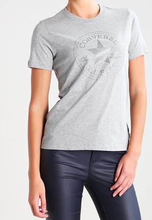 Personne portant un t-shirt gris Converse All Star et un pantalon ajusté bleu foncé, avec une main posée sur la hanche.