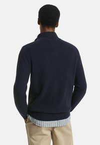 Marineblauer Strickpullover mit hohem Kragen und gerippten Bündchen und Saum. Über einem hellblau gestreiften Hemd getragen. Weiche Textur, lässiges Design.