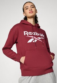 Felpa bordeaux realizzata in morbido tessuto, con tasca frontale, caratterizzata da un logo Reebok bianco e cappuccio con cordino. Maniche lunghe con polsini a costine.