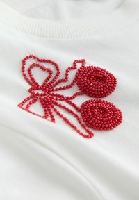 Valge rõivaeseme punase helmestega tikitud kirsi ja paela mustriga, millel on kaks helmestest kirssi ja pael, esitledes sujuvat tekstuuri ja elavat detaili.