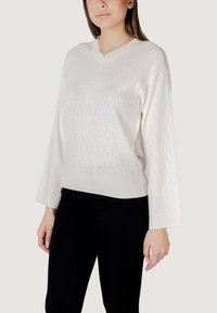 Maglione bianco a maglia con scollatura a V, caratterizzato da un motivo geometrico e maniche testurizzate, abbinato a jeans neri.