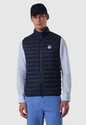 Giovane uomo che indossa un gilet trapuntato blu navy sopra una camicia a righe chiare, pantaloni blu e cappello blu navy, rivolto in avanti con espressione neutra.