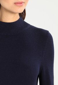 Dunkelblauer Strickpullover mit einem gerippten Stehkragen, langen Ärmeln und subtiler Textur, der eine enganliegende Silhouette aufweist.