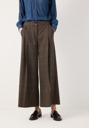 Person trägt braune karierte, weit geschnittene Hose, blaues Langarmhemd mit Knöpfen und dunkelbraune Loafer, steht mit den Händen in den Taschen.