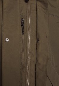 Gros plan d'une veste marron montrant une fermeture éclair, une petite poche zippée, et deux boutons-pression en métal sur le tissu avant.