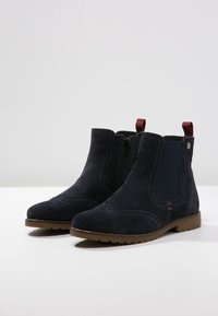 Bottes chelsea en daim marine foncé avec des panneaux élastiques sur les côtés, des coutures décoratives et une semelle en caoutchouc marron. Elles ont un petit tirant rouge à l'arrière.