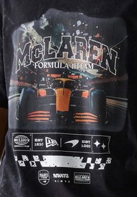 Czarna koszulka z żywą grafiką samochodu Formuły 1 z pomarańczowymi akcentami. Zawiera napis "Zespół Formuły 1 McLaren" oraz różne logo.