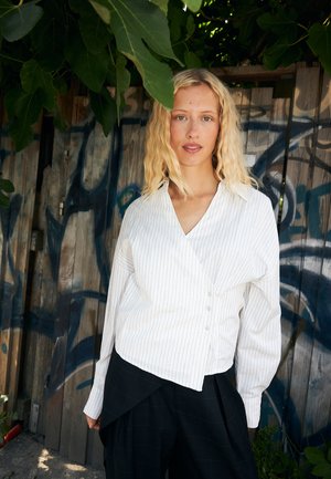 Femme blonde portant un chemisier cache-cœur blanc à fines rayures et un pantalon noir, debout à l'extérieur devant une clôture en bois avec des graffitis et des feuilles vertes au-dessus.