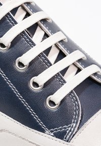 Gros plan sur une basket bleu marine et blanche avec des lacets blancs passant à travers des œillets métalliques et des détails de couture visibles.