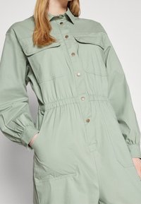 Mono utilitario de algodón verde con mangas largas, bolsillos en el pecho y cintura elástica. Presenta botones a presión y un corte holgado.