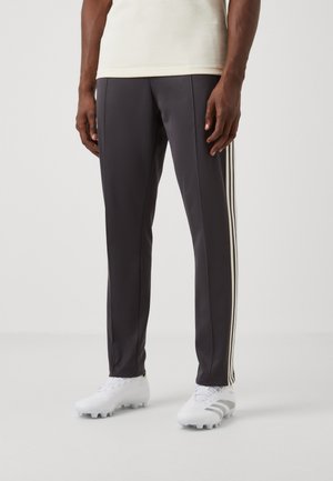 Schwarze Sporthose mit weißen Seitenstreifen, getragen von einer stehenden Person, kombiniert mit weißen Fußballschuhen und einem hellbeigen Oberteil.