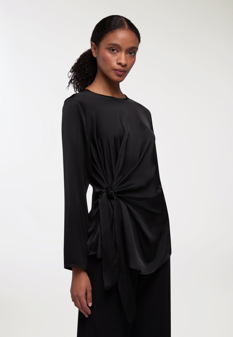 Top in satin nero con maniche lunghe, caratterizzato da un dettaglio frontale attorcigliato legato in vita, texture liscia e scollatura rotonda.