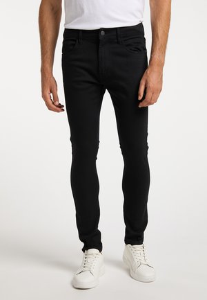Mann mit schwarzen Skinny Jeans und weißen Sneakers steht vor einem einfarbigen Hintergrund.