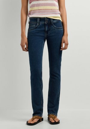 Vrouw draagt donkerblauwe rechte jeans, een gestreept mouwloos topje en bruine sandalen, staand tegen een effen achtergrond.