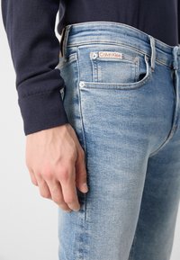 Jeans de mezclilla azul claro con un diseño ajustado, con una etiqueta del logo de Calvin Klein, costuras visibles y un ligero desgaste en la tela.