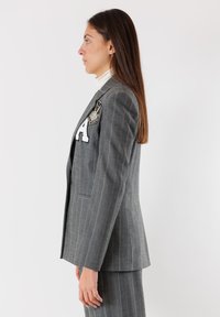 Blazer grigio a righe con una vestibilità strutturata, caratterizzato da una patch con lettere e un emblema decorativo sul lato sinistro. Tessuto liscio con un aspetto sartoriale.