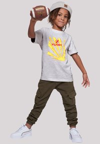 Camiseta gris con gráfico de "FLASH", diseño de explosión amarilla, pantalones cargo oliva y zapatillas blancas. Niño sostiene un balón de fútbol marrón.