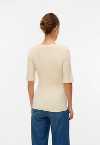 Top a maniche corte a coste in beige chiaro; design aderente con scollatura tonda e texture liscia, abbinato a jeans blu larghi.