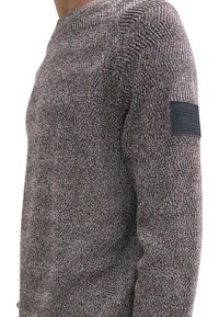 Pull en texture avec un motif en chevrons gris, rouge et bleu. Présente une étiquette noire sur la manche gauche, un col rond et des manches longues.