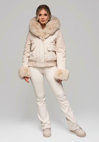 Beżowa kurtka puchowa z dużym sztucznym futrzanym kołnierzem i pasującymi mankietami, zestawiona z dopasowanymi kremowymi spodniami i beżowymi sneakersami na platformie.