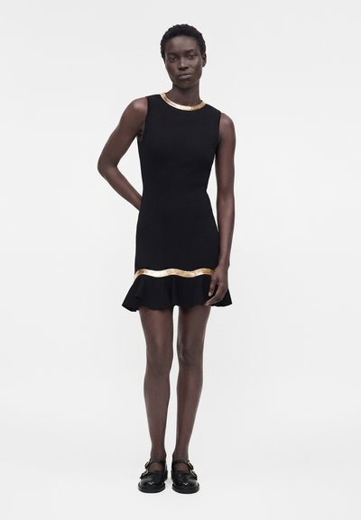 Γυναίκα που στέκεται, φοράει μαύρο αμάνικο φόρεμα με χρυσή επένδυση στον λαιμό και πάνω από το βολάν στο τελείωμα, σε συνδυασμό με μαύρα μοκασίνια.