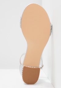 Silver strappy sandal with a smooth sole, textured braided accents, and a light brown rubber outsole, featuring minimal design elements.