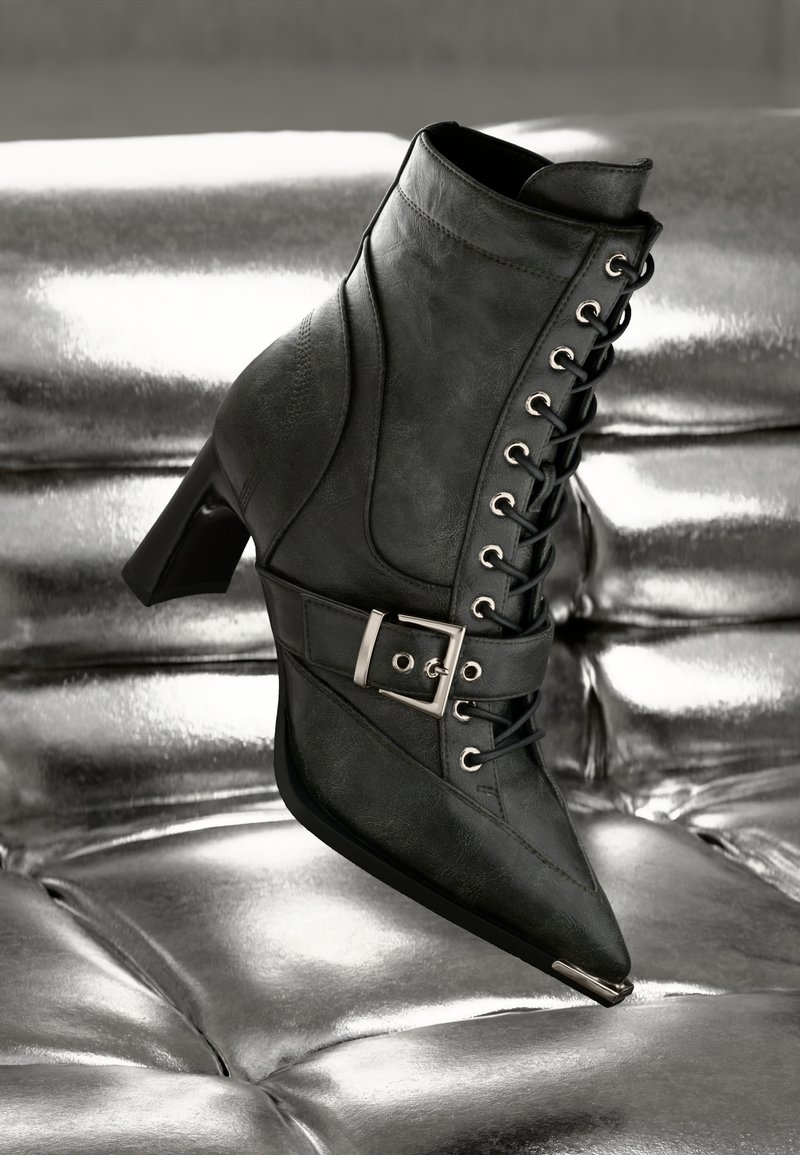 Musta nahkasaapas nilkkaremmillä, terävä kärki, nauhakiinnitys, koristeellinen solki ja stiletto-korko, asetettuna hopean teksturoidulle taustalle.