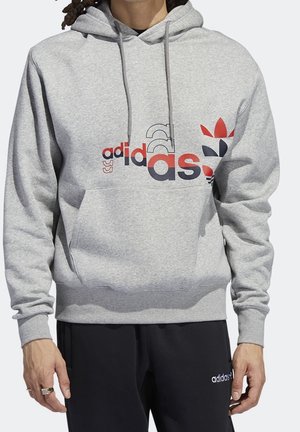 Person iført en grå Adidas hættetrøje med flerfarvet logo og sorte Adidas bukser med hvid logo tekst på højre lår.