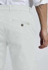 Pantalones de algodón verde menta claros con corte recto, que cuentan con un bolsillo trasero con cierre de botón y un detalle de lazo de tela.