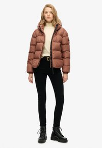 Chaqueta de plumas marrón con capucha, diseño acolchado, cremallera frontal y bolsillos laterales, llevada sobre un suéter de cuello alto color crema y jeans negros. Botas negras de cordones.