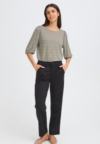 Pantalon noir à coupe ajustée, associé à un blouse verte texturée avec un motif à carreaux et des manches bouffantes. Le modèle se tient avec les mains dans les poches.