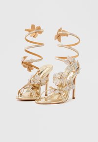 Kullast kõrge kontsaga sandaalid, millel on lillelised kristallkaunistused, spiraalsed seemisnahast pahkluu rihmad ja avatud varbaga disain.