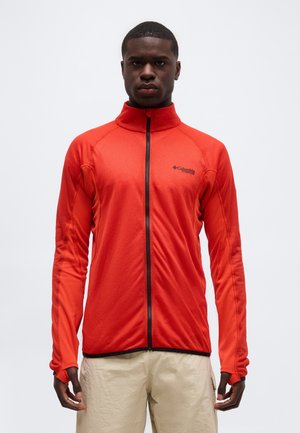 Homme portant une veste de sport zippée Columbia orange vif avec des trous pour les pouces, associée à un pantalon beige clair, faisant face à l'avant sur un fond uni.