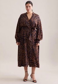 Abito midi a stampa leopardata in marrone e nero. Maniche lunghe, cintura con fiocco e chiusura con bottoni sul davanti. Tessuto liscio e vestibilità comoda. Indossato con tacchi neri.