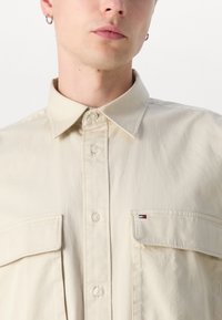 Beige overhemd met knopen en een klassieke kraag, voorzien van twee voorzakken en een kleine geborduurde logo patch op de linkerborstzak.