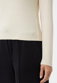 Jasnobeżowa bluzka z długim rękawem o dopasowanym kroju i miękkiej fakturze. Posiada subtelne logo przy ściągaczu, zestawiona z ciemnymi, czarnymi spodniami.